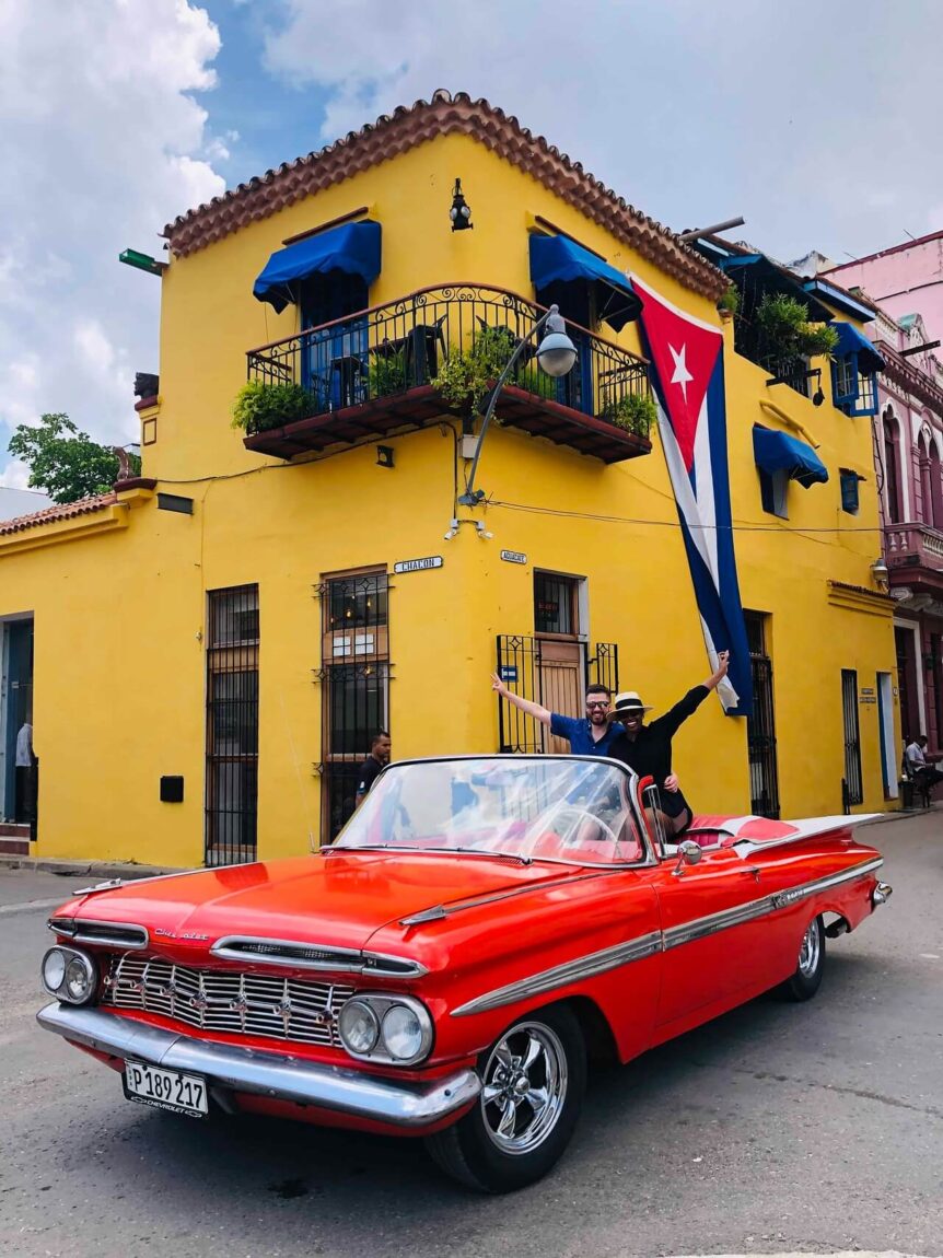 Tourists enjoying a ride in a red classic convertible car in Havana, Cuba, with a yellow colonial building and the Cuban flag in the background.